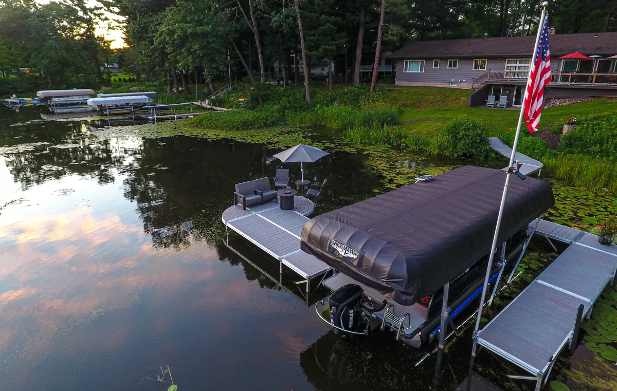 An aerial view of a waterfront property featuring a modular dock system, a boat lift with a black canopy cover and outdoor seating with an umbrella.